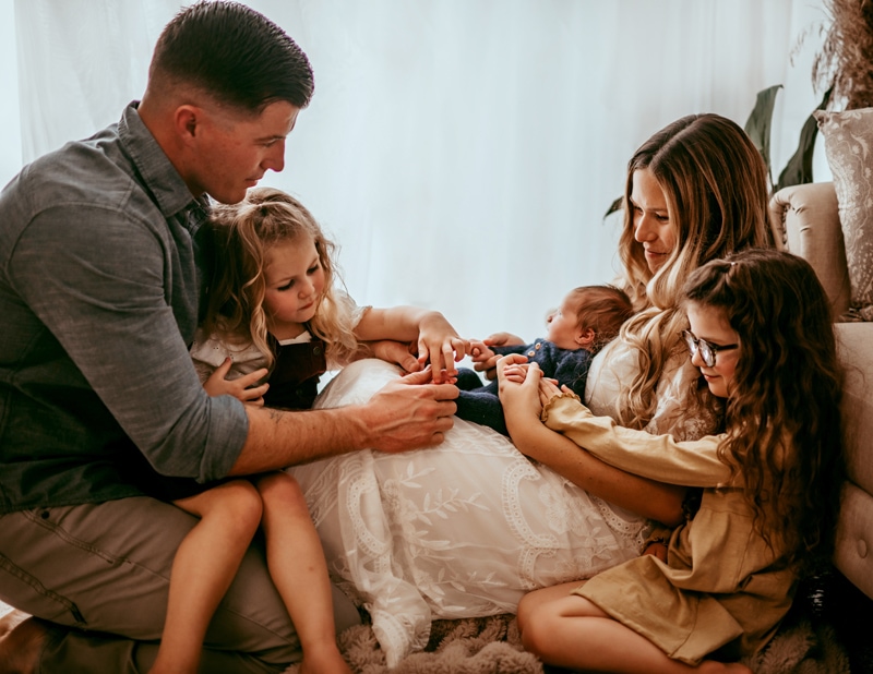 Family Photographer, Mom, dad, and two daughters all gather around baby brother at home