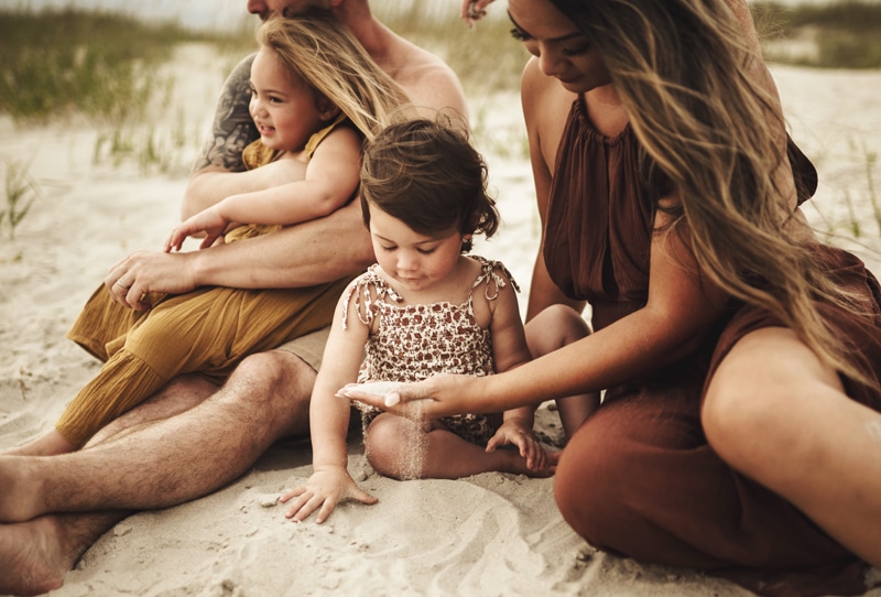 Family Photographer, Mom and dad sit on a sandy beach with their two young daughters